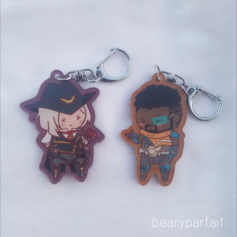 May include: Two character keychains featuring anime-style illustrations. The keychain on the left features a female character with white hair, wearing a brown hat and a black and gold outfit. The keychain on the right features a male character with brown skin, black hair, and a blue and orange outfit. Both keychains have silver key rings.
