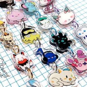 May include: A collection of clear acrylic keychains featuring various cartoon characters. The keychains have gold-colored metal rings and colorful designs, including a banana, octopus, cat, and bear. The background is a grid pattern.