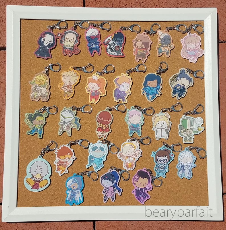 May include: A corkboard with a variety of colorful keychains featuring cartoon characters. The keychains are attached to the corkboard with metal rings. The corkboard is framed in white and has the text "bearyparfait" written on the bottom right corner.