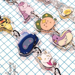 以下が含まれることがあります： パステルカラーの様々な漫画のキャラクターが描かれた、透明なアクリルキーホルダーのコレクション。各チャームは小さな黒いリングとシルバーの留め金で接続されています。キーホルダーは青と白のグリッドの背景にあります。