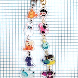 May include: A keychain featuring multiple clear acrylic charms with colorful cartoon character designs. The charms are connected by small metal rings and a gold-colored clasp. The characters include an orange creature, a black and white cat, and a blue and white robot.