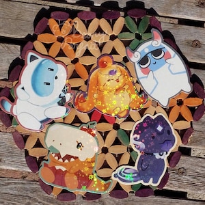 May include: A collection of five colorful cartoon character stickers, including a white cat-like creature, a yellow bunny, a blue and white ghost, a dinosaur, and a purple skunk. The stickers are arranged on a decorative wooden coaster with a floral pattern.