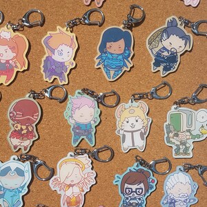 May include: A collection of 16 Overwatch character keychains. The keychains are made of clear acrylic and feature various characters from the game, including Tracer, D.Va, Genji, and Mercy. Each keychain has a silver metal ring for attaching to keys or bags.