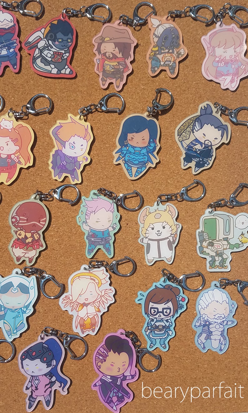 May include: A collection of 16 Overwatch character keychains. The keychains are made of clear acrylic and feature various characters from the game, including Tracer, D.Va, Genji, and Mercy. Each keychain has a silver metal ring for attaching to keys or bags.
