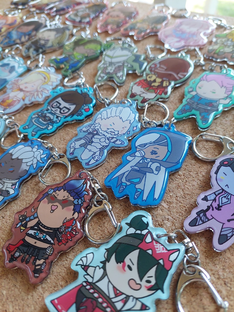 May include: A collection of colorful character keychains featuring various anime and video game characters. The keychains are made of clear acrylic and have a glossy finish. Some of the characters include a girl with blue hair and a red dress, a girl with pink hair and a white dress, and a boy with white hair and a blue suit.