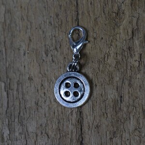 May include: Silver charm with a button design. The button has four holes and is attached to a silver jump ring.