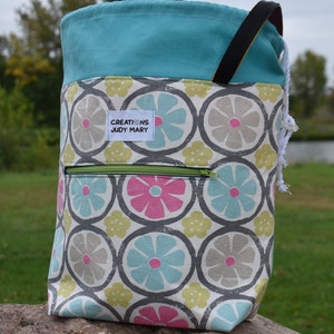 May include: A turquoise and white drawstring bag with a black handle and a zippered pocket. The bag has a floral print with pink, blue, and yellow flowers. The bag has a label that says "Creations Judy Mary".