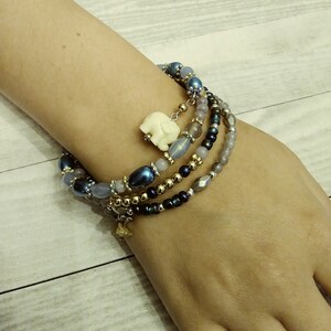 Op de afbeelding: Een meerstrengs armband met blauwe, grijze en gouden kralen. Een kleine witte olifant bedel hangt aan de armband.