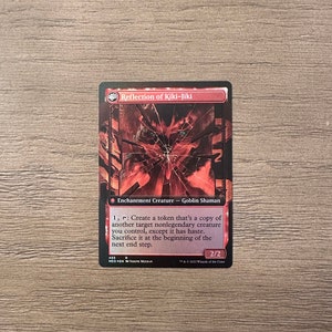 May include: A red and black Magic: The Gathering card with the text "Reflection of Kiki-Jiki" and the text "Enchantment Creature - Goblin Shaman". The card has a red and black illustration of a goblin with a cracked face.
