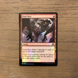 May include: A red and black Magic: The Gathering card with the title "Dual Shot". The card is an instant and deals 1 damage to each of up to two target creatures. The card art features a woman with a gun and a red background.