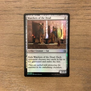 May include: A Magic: The Gathering card featuring a white background with a silver border. The card depicts two cat-like creatures with glowing eyes standing in front of a stone structure. The card's name is "Watchers of the Dead" and the text reads "Exile Watchers of the Dead: Each opponent chooses two cards in his or her graveyard and exiles the rest. They are tasked with protecting the entrance to the embalming chambers."