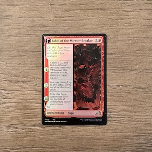 May include: A red and black Magic: The Gathering card with the title "Fable of the Mirror-Breaker". The card is a 2/2 enchantment - saga with three abilities. The card art depicts a dark, cracked, and broken mirror.