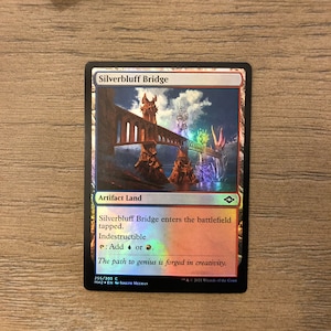May include: A Magic: The Gathering card depicting a multi-colored bridge with a fantasy landscape. The card is titled "Silverbluff Bridge" and is an Artifact Land. The card text reads: "Silverbluff Bridge enters the battlefield tapped. Indestructible. Add or. The path to genius is forged in creativity."