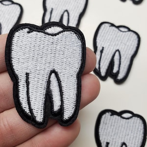 May include: A collection of embroidered patches shaped like teeth. Each patch features a white tooth with black outlines and a black border. The patches are designed for decorative purposes and can be applied to clothing or accessories.