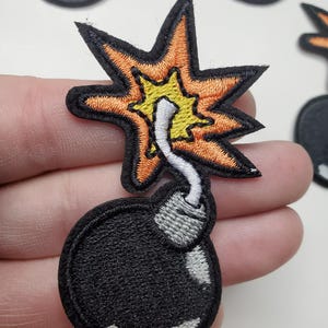 May include: Embroidered patch featuring a black bomb with a gray fuse and a starburst explosion in orange and yellow. The patch has a black border and is likely for clothing or accessories.