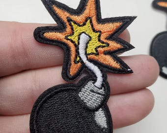 May include: Embroidered patch featuring a black bomb with a gray fuse and a starburst explosion in orange and yellow. The patch has a black border and is likely for clothing or accessories.