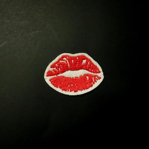 May include: Embroidered patch featuring a pair of red lips with white accents. The lips are detailed with visible stitching and are set against a white border. The patch is likely designed to be ironed or sewn onto fabric.