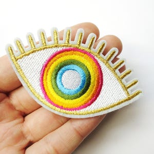 May include: Embroidered eye patch with a white base, gold outline, and a rainbow-colored iris. The patch has a white eyelash design and is likely for clothing or accessories.