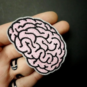 May include: A pink brain patch with black embroidered details and a white border. The patch is designed to resemble a human brain and is likely intended for decorative purposes, such as on clothing or accessories.
