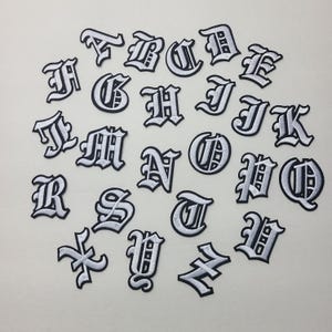 May include: A set of 26 white iron-on letters in an old English font style. The letters are arranged in alphabetical order.
