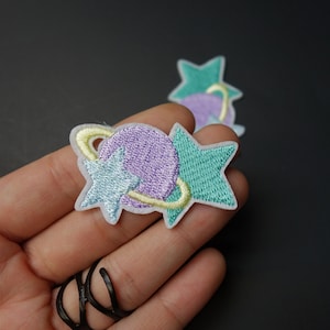 May include: Embroidered patches featuring a purple planet with yellow rings and two stars in shades of blue and green. The patches have a white border and are likely for clothing or accessories.