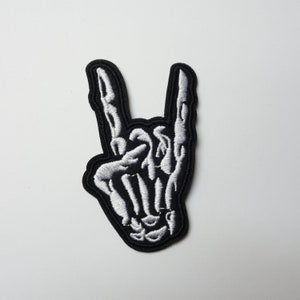 May include: Embroidered patch featuring a white skeleton hand in the 'rock on' gesture, set against a black background. The patch has a black border and is designed for application on clothing or accessories. The design is detailed and the stitching is precise.