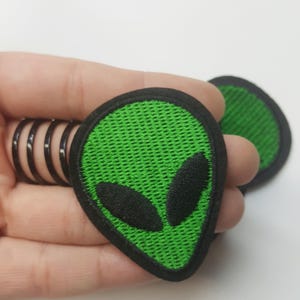 May include: Two green alien head embroidered iron-on patches with black outlines. The patches have a textured green fabric and black eyes.