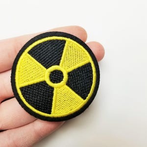 May include: Embroidered patch featuring a black and yellow radiation symbol. The circular patch has a black border and a central design of alternating black and yellow segments, with a yellow circle in the center. The patch is likely for clothing or accessories.