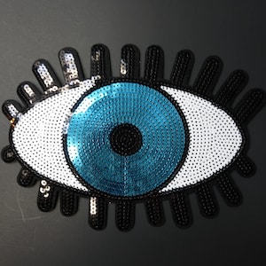 May include: A decorative eye-shaped patch with a blue iris, white sclera, and black eyelashes. The iris is a vibrant blue, surrounded by a black outline. The sclera is white with small, reflective sequins. The eyelashes are black.