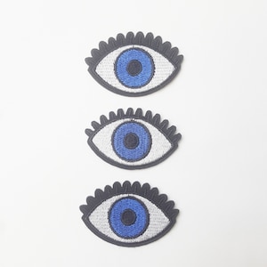 May include: Three embroidered eye patches. Each patch features a blue iris, a white sclera, and a black, scalloped edge. The patches are arranged vertically against a white background.