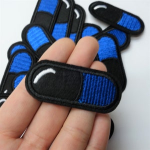 May include: A set of blue and black embroidered iron-on patches shaped like pills. The patches have a white outline and a white line across the middle.