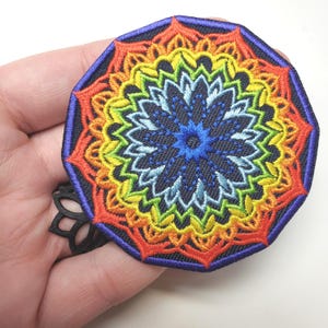 May include: A colorful embroidered patch featuring a mandala design. The patch has a dark blue background with a vibrant rainbow of colors, including red, orange, yellow, green, and blue. The patch is octagonal with a dark blue border.