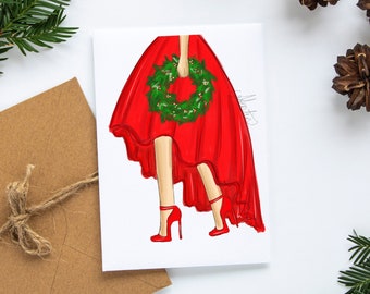 May include: A Christmas-themed greeting card featuring a fashion illustration of a person in a red dress holding a green wreath. The card is on a white surface, with a brown envelope and pine needles in the background.