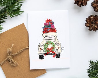 May include: A white greeting card with a festive illustration of a vintage truck carrying a stack of wrapped gifts. The truck has a wreath and the word "Holidays" on the front. A brown envelope and pine needles are in the background.