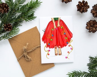 May include: A Christmas-themed greeting card featuring a fashion illustration of a person holding a red skirt adorned with colorful Christmas lights. The card is accompanied by a brown envelope and festive greenery.