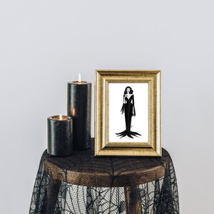 May include: A gold-framed illustration of a woman in a black dress, displayed on a small table. Two black candles with lit flames are next to the frame. The table is covered with a black lace cloth.