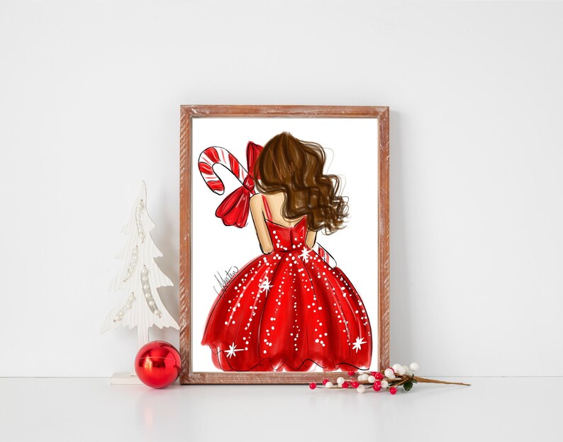 Op de afbeelding: Een ingelijste illustratie van een vrouw in een rode jurk die een zuurstok vasthoudt. De jurk is versierd met witte stippen. Het kunstwerk heeft een kerstthema en wordt op een wit oppervlak gepresenteerd.