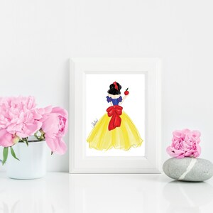 May include: A framed art print featuring a watercolor illustration of a woman in a yellow dress with a red bow and holding an apple. The artwork is in a white frame, with pink flowers in a white mug and on a stone.
