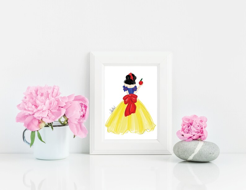 May include: A framed art print featuring a watercolor illustration of a woman in a yellow dress with a red bow and holding an apple. The artwork is in a white frame, with pink flowers in a white mug and on a stone.