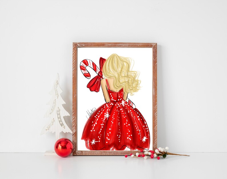 Op de afbeelding: Een ingelijste print met een mode-illustratie van een vrouw in een rode jurk met witte sneeuwvlokaccenten, die een zuurstok vasthoudt. De vrouw heeft blond haar en een rode strik. Het kunstwerk staat tegen een witte achtergrond.