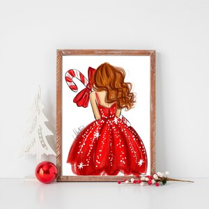 Op de afbeelding: Een ingelijste print met een mode-illustratie van een vrouw in een rode jurk met witte stippen, die een zuurstok vasthoudt. Het kunstwerk heeft een kerstthema, met een rode strik en de handtekening van de kunstenaar.