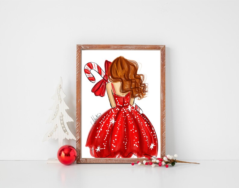 Op de afbeelding: Een ingelijste print met een mode-illustratie van een vrouw in een rode jurk met witte stippen, die een zuurstok vasthoudt. Het kunstwerk heeft een kerstthema, met een rode strik en de handtekening van de kunstenaar.
