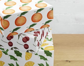 May include: Three stacked gift boxes with white backgrounds and fruit patterns. The top box features oranges with green leaves. The middle box has cherries. The bottom box has lemon slices and green leaves. The boxes are on a wooden surface.