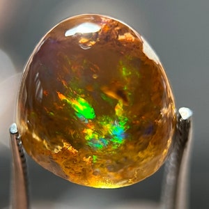 Puede incluir: Una piedra preciosa de ópalo de color marrón dorado con destellos iridiscentes verdes y azules. El ópalo tiene forma ovalada y una superficie lisa y pulida.