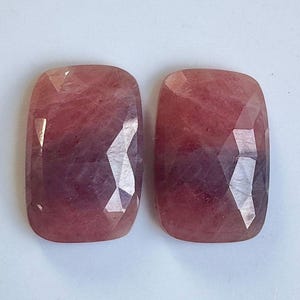 May include: Two faceted, rectangular gemstones with rounded corners. The stones display a gradient of pink and purple hues, with a polished, reflective surface. The gemstones are likely for jewelry making.
