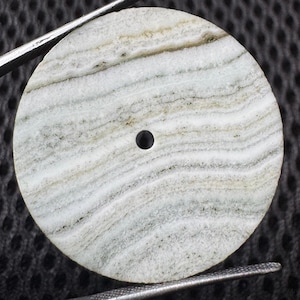 May include: A circular, flat stone pendant featuring a natural banded pattern of white, grey, and light brown layers. The stone has a smooth, polished surface and a small, centered circular hole for threading, held by metal tweezers.