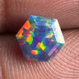 May include: A faceted, hexagonal opal gemstone. The opal displays a dark base color with vibrant flashes of orange, yellow, green, and blue. The gemstone is held in a hand, showcasing its intricate internal play of color.