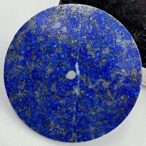 May include: A circular lapis lazuli stone, displaying a rich blue hue with gold inclusions. It features a small, drilled hole. The stone measures approximately 7.6 cm in diameter, ideal for jewellery or craft projects. The surface is polished.
