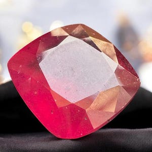 Puede incluir: Una gema facetada, de talla cojín, con un vibrante tono rojo. La superficie de la gema refleja la luz, mostrando sus múltiples facetas y claridad interna. La gema se encuentra sobre un fondo oscuro, resaltando su color y brillo. Podría ser un rubí o una piedra preciosa similar.