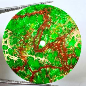 Puede incluir: Un disco de piedra preciosa plano y circular que presenta una base de color verde vibrante con vetas intrincadas en forma de red en tonos rojo oscuro y bronceado claro. La pieza tiene un pequeño orificio circular centrado y está sujeta por pinzas.